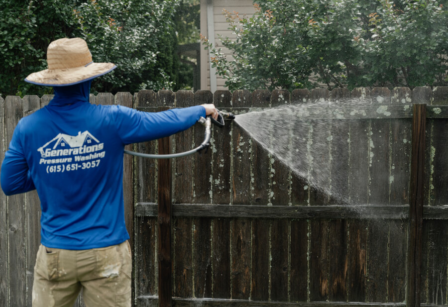 A person seen from behind is using a pressure washer to clean a dirty, dark wood fence covered with patches of lichen.