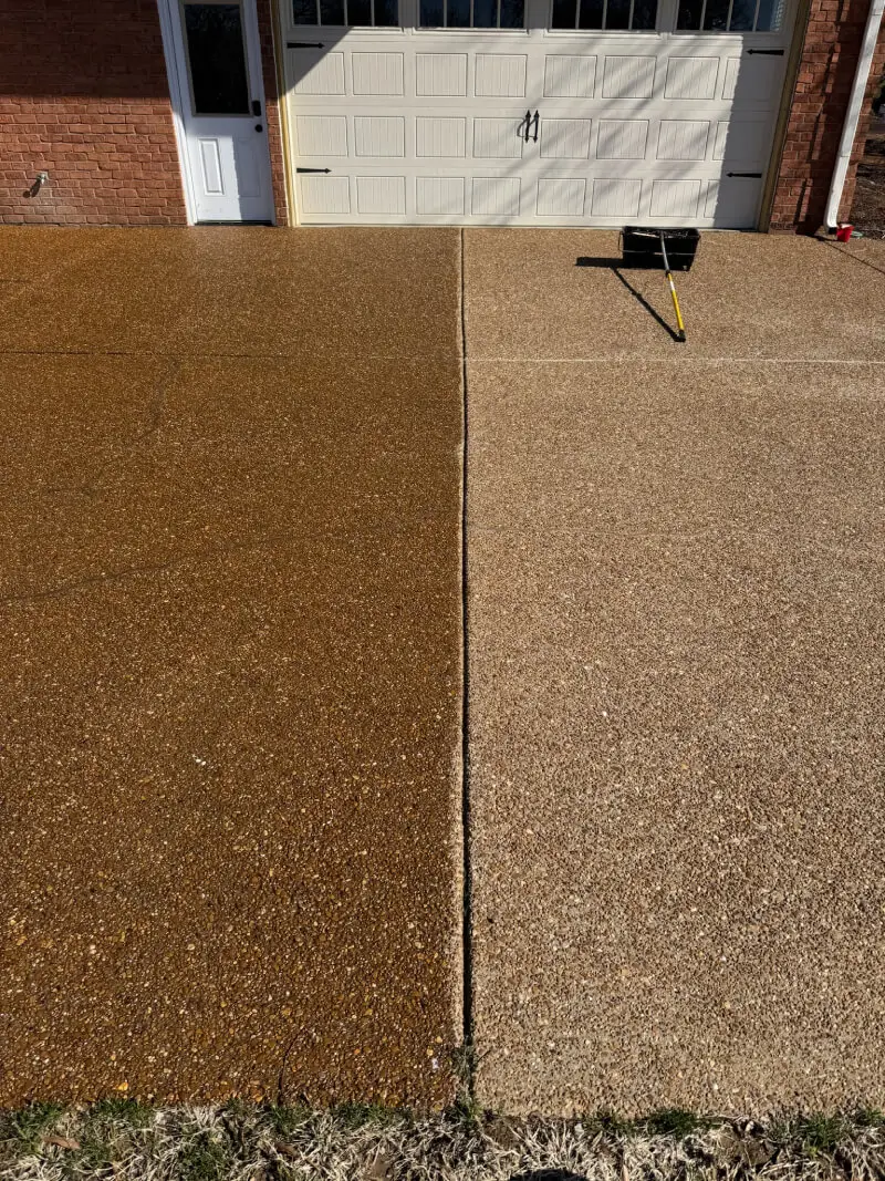 A detailed photograph showing a striking before-and-after contrast on an exposed aggregate concrete driveway surface, where the left section is dark, rich, and enhanced by a newly applied sealer, while the right section remains lighter and duller. A long-handled application roller lies resting on the unsealed right side near the base of a brick house.