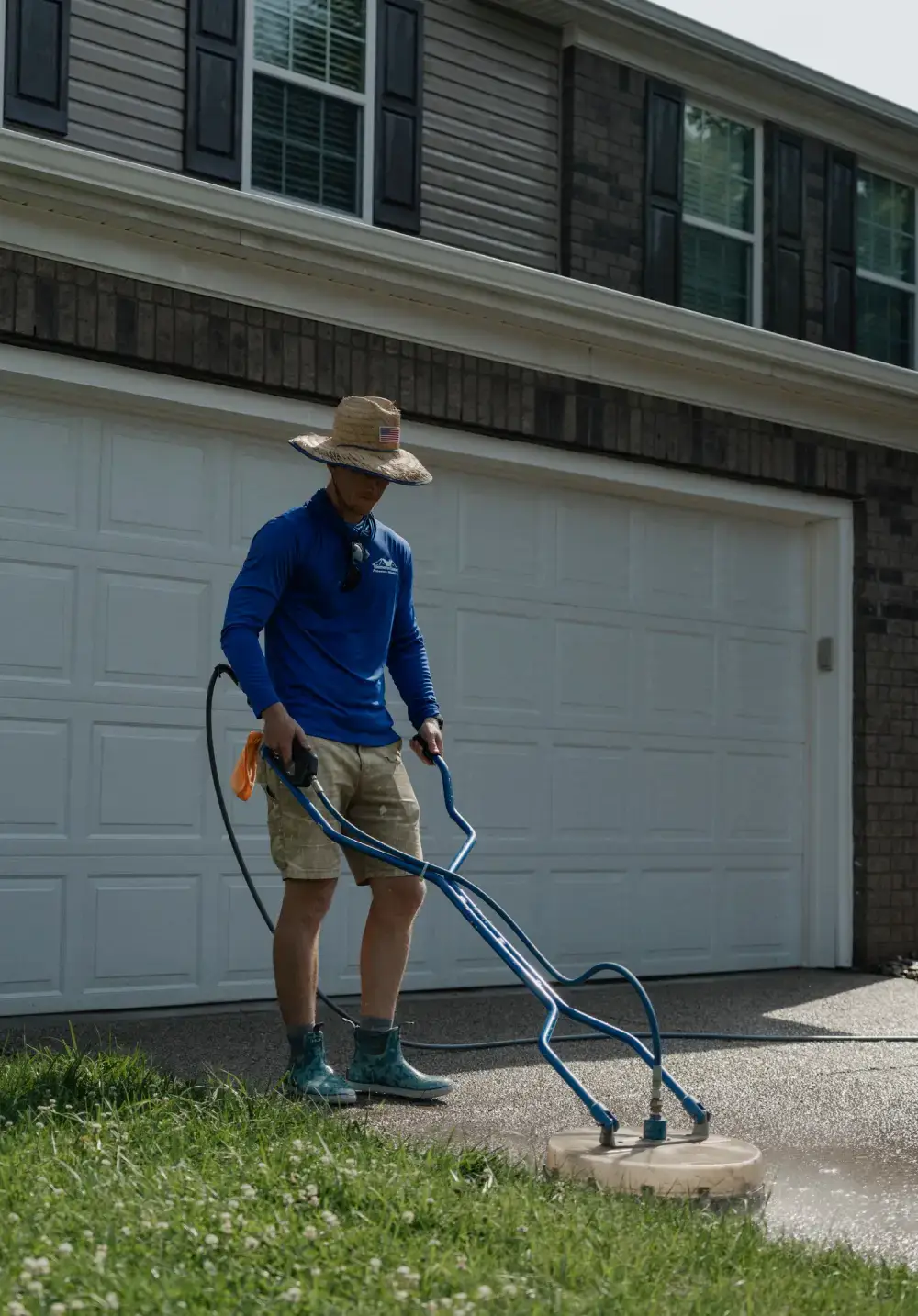 A pressure washing professional wearing a straw hat and blue uniform shirt uses a blue surface cleaner on the concrete driveway of a two-story brick and siding home.