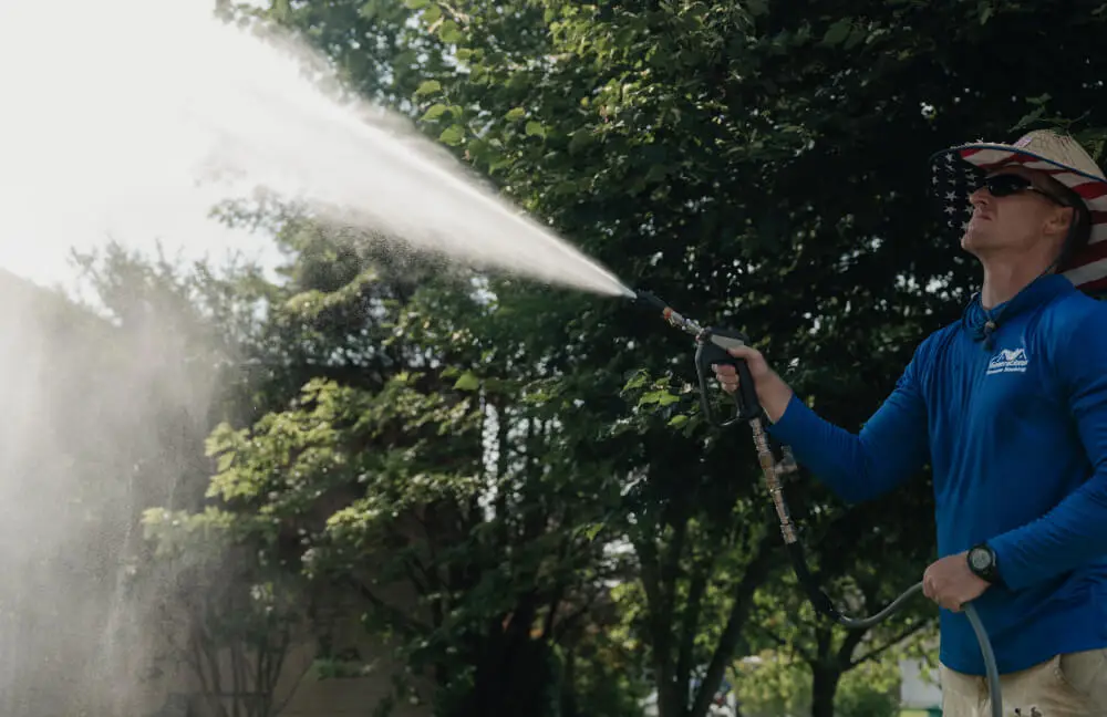 A side-profile view shows a pressure washing professional wearing sunglasses and a wide-brimmed straw hat with an American flag pattern underneath the brim. He is dressed in a blue long-sleeved shirt and is using a spray gun to mist water upward toward lush green trees, with bright sunlight filtering through.